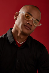 Wall Mural - Close-up shot of a man with shaved head in a black shirt over a red collar and with cat eyes retro sunglasses with a transparent half-frame and yellow lens. The photo is made on the red background.