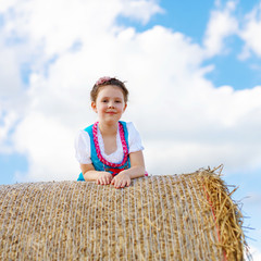 Wall Mural - Cute little kid girl in traditional Bavarian costume in wheat field. German child with hay bale during Oktoberfest in Munich. Preschool girl play at hay bales during summer harvest time in Germany.