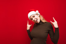 Joyful Girl In Santa Hat Listens To Rock Music In Wireless Headphones On Red Background, Looks Into Camera, Smiles And Shows Hands With Heavy Metal Gesture. Xmas