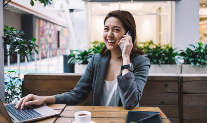 Cheerful adult beautiful freelance female having phone call while typing on laptop at cafe