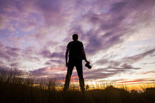 Rear View Of A Young Man Standing With His Photo Camera And Watching A Colourful Sunset With Purple Clouds