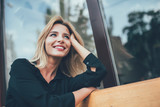 Fototapeta  - Positive cheerful hipster girl with perfect blonde hair taking rest on bench in city and enjoying time for dreaming outdoors, happy female with cute candid smile looking away during leisure