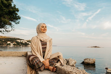 Portrait Of Young European Muslim Women With Hijab Sitting On The Stone Beach With Sea In The Background. She Is Happy And Relaxed.
