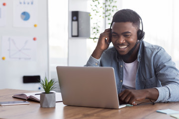 Black worker wearing headphones, watching webinar on laptop in office