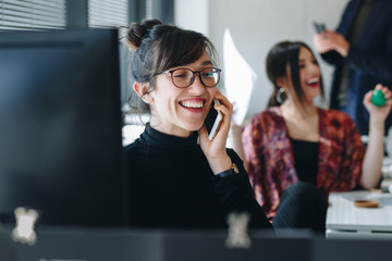 Portrait of happy businesswoman talking on the phone in creative office. Coworkers in the background. Happy and busy office life concept.