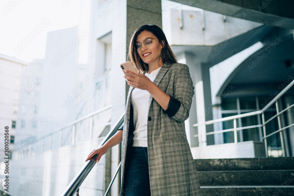 Successful female entrepreneur dressed in formal jacket enjoying online communication on website typing sms answer via smartphone gadget, positive young woman using 4g wireless on cellphone - obrazy, fototapety, plakaty 