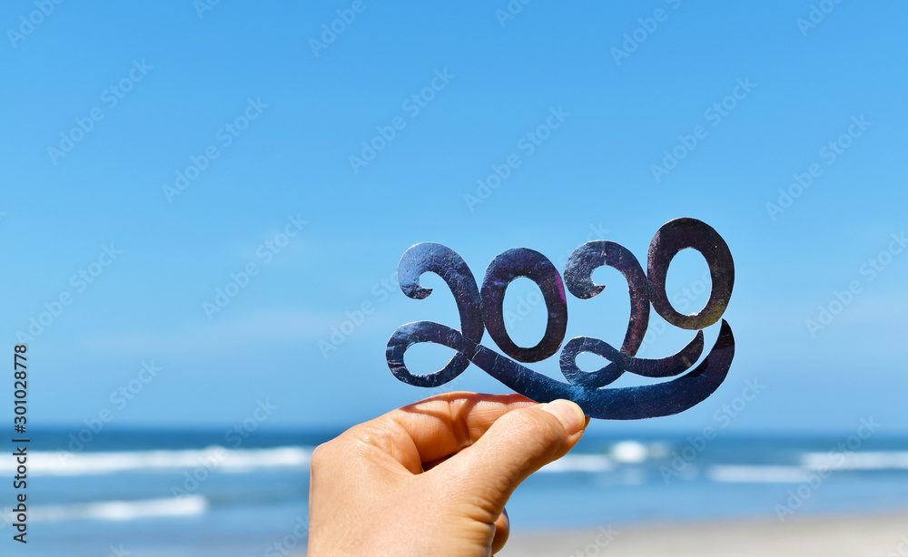 Happy New Year creative concept, hand holding 2020 text against blue sky and beach blur defocused background. - obrazy, fototapety, plakaty 
