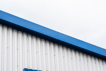 Metal corrugated sheets on a building with a blue metal corners. White aluminium metal corrugated roof or wall sheets against cloudy sky background on a factories and industrial buildings.