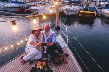 Happy senior couple taking selfie photo at night time on a sailboat, during anniversary vacation - Focus on hands phone