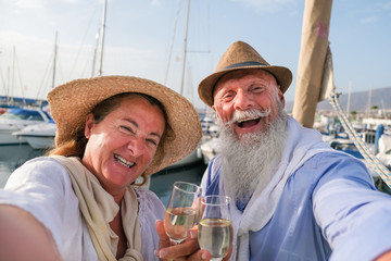 Wall Mural - Happy senior couple doing selfie, cheering with champagne on a sailboat during anniversary vacation - Joyful elderly lifestyle, love, travel and holidays concept - Focus on faces