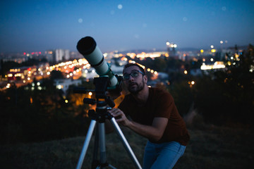 Wall Mural - astronomer with a telescope watching at the stars and moon with blurred city lights in the backgroun