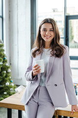 happy businesswoman holding cup near decorated christmas tree