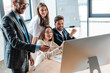 © LIGHTFIELD STUDIOS - happy businessman in glasses holding pen and looking at computer monitor near multicultural coworkers in office