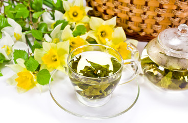 Wall Mural - Green tea in a glass cup with tea leaves,  daffodil flowers and green tree branches, wicker basket on a white background. Spring still life.