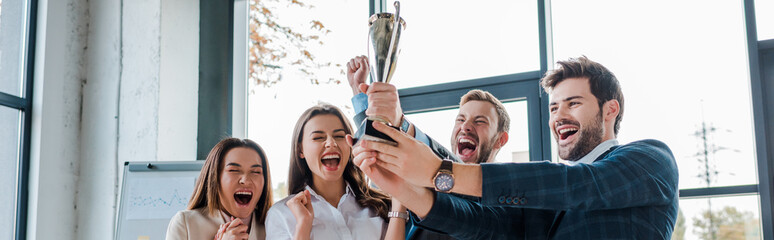 Wall Mural - panoramic shot of excited multicultural businesswomen and businessmen looking at trophy in office