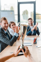 cropped view of businesswoman holding champion cup near coworkers in office