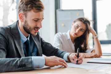 Wall Mural - selective focus of sad businessman holding pen near paper and attractive businesswoman in office