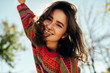© iuricazac - Bottom view close-up portrait of happy young woman smiling broadly with windy hair and freckles has joyful expression, wearing colorful knitted sweater with arms wide open, posing on nature sunlight