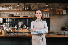 Smiling Barista In Apron Standing With Crossed Arms Near Bar Counter