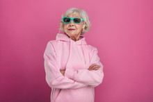 Cool Old Woman In Funny Green Sunglasses Posing On Camera. Hands Crossed. Wear Pink Hoody. Stylish Modern Grandma. Isolated Over Pink Background.