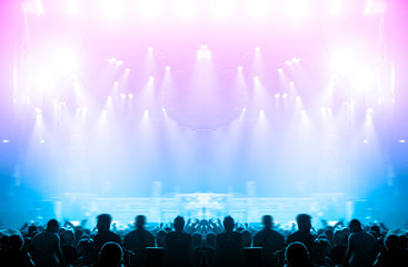 Wall Mural - Shot taken inside a concert hall, in the middle of the crowd. The main stage is visible, fully lit in the night; people silhouettes are visible, clapping, dancing or holding smartphones for photo.