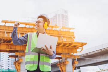 Wall Mural - Image outside the industrial construction engineers in yellow protective ear muff discuss new project while using laptop and happy smile on the open building site near the crane.