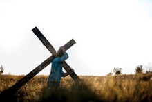 Male Carrying A Handmade Wooden Cross On His Back In A Grassy Field