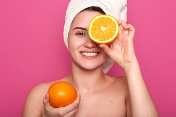 Indoor short of young female holding orange in one hand and covering her eye with half of fruit, winsome lady with pefect skin, posing isolated over pink background. Woman skin care concept.