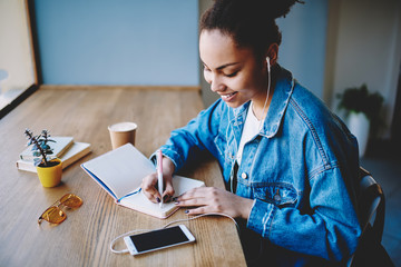 Wall Mural - Positive dark skinned female student writing essay with excited idea while enjoying favourite music playlist with motivation mood, happy meloman making notes of name of songs in textbook for education