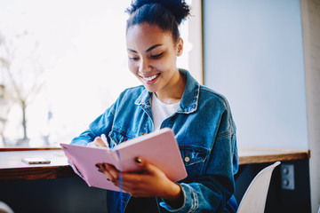 Wall Mural - Cheerful dark skinned female student writing positive article in textbook during free time in cafeteria, happy hipster girl on trendy jeans jacket planning course work with knowledge notebook