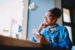 © BullRun - Happy hipster girl in stylish spectacles spending time with smartphone device in cafeteria connected to wireless 4g internet, positive woman dreaming about future life while holding telephone in hand