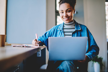 Wall Mural - Portrait of cheerful female student sitting in cafeteria and smiling at camera during break from exam preparation, happy hipster girl research information for college course work using laptop device