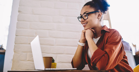 Wall Mural - Happy black woman in optical eyewear for vision correction reading information in profile of famous blogger in social networks, cheerful hipster girl watching positive movie on content website