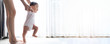 Asian baby taking first steps walk forward on the mat. Happy little baby learning to walk with mother help at home. Mother teaching how to walk gently. Baby growth and development concept. Banner size