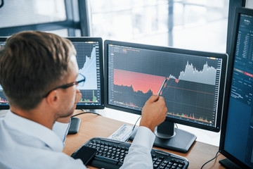 Looks at the graphs. Young businessman in formal clothes is in office with multiple screens. Conception of exchange and money