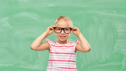 Wall Mural - education, school and vision concept - smiling cute little girl in black glasses over green chalkboard background