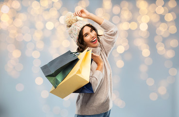 Sticker - christmas, seasonal sale and consumerism concept - happy smiling young woman in knitted winter hat and sweater with shopping bags over festive lights background