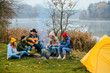 © Iryna - Hipster man playing on acoustic guitar and singing song with friends travelers at big bonfire at night camp in the forest. Group of people chilling at fire in the evening, camping near lake