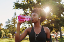 Fitness Athlete Young African American Woman Listening To Music On Earphones Drinking Water In A Reusable Water Bottle After Working Out Exercising On Sunny Day At The Park