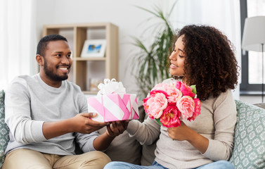 Wall Mural - holiday, greeting and people concept - happy african american couple with bunch of flowers and gift at home