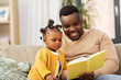 © Syda Productions - family, fatherhood and people concept - happy african american father reading book for baby daughter at home
