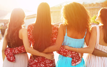 Calm Women Relaxation. Four Beautiful Girls Are Posing With Their Backs To The Camera In Multicolored Dresses, Hugging Each Other Like Best Friends.