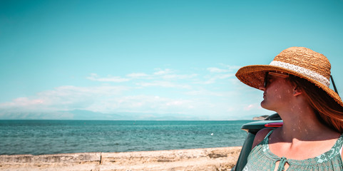 Poster - Young woman in a straw hat and sunglasses. Dark soul car on the beach. View of beautiful sea and blue sky. Place for your text or product. A beautiful summer day. Car trip and sightseeing of the coast