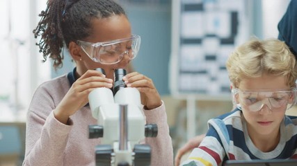 Poster - Elementary School Science Classroom: Cute Little Girl Looks Under Microscope, Boy Uses Digital Tablet Computer to Check Information on the Internet. Teacher Observes from Behind