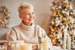 © Anatoliy Karlyuk - Xmas, holidays, decor, party and festive atmosphere concept. Good looking cheerful middle aged female with short hair enjoying Christmas mood, sitting around wax candles, decorations and lights