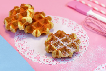 Wall Mural - Fresh small round waffles with sugar powder on paper lace doily and pink ground with baking utensils in background