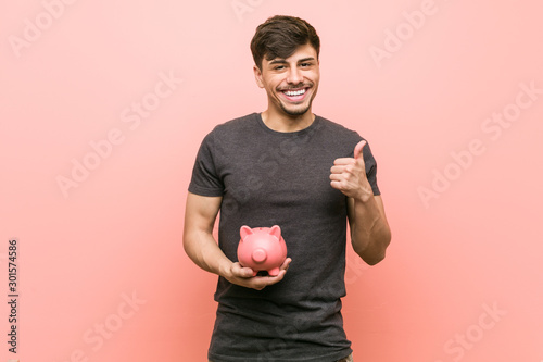 Fotografía Young hispanic man holding piggy bank smiling and raising thumb up