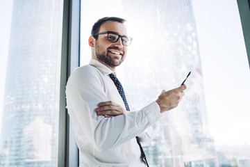 Wall Mural - Half length portrait of cheerful male proud ceo feeling good during working day in company holding cellular gadget and smiling at camera, happy man in optical spectacles for provide eyes protection