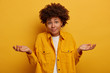 Puzzled clueless African American lady shrugs shoulders and expresses uncertainty, makes decision, wears fashionable clothing, spreads palms sideways isolated on yellow wall. Life perception, attitude