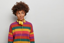 Photo Of Thoughtful Curly Haired Woman Looks Aside With Pensive Expression, Wears Headphones And Striped Jumper, Has Plan In Mind, Poses Over White Background, Blank Space For Your Advertising Content
